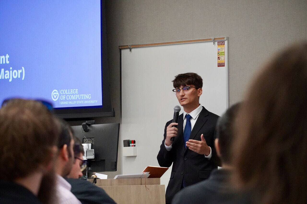A student in a dark suit, blue tie, and glasses speaks into a microphone during a presentation at Grand Valley State University’s College of Computing. The presentation slide beside him displays the college’s logo and title while audience members l...